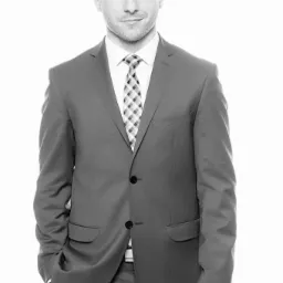 Portret młodego mężczyzny w garniturze i krawacie w kratę na jasnym tle, z ręką w kieszeni, uśmiechający się lekko.