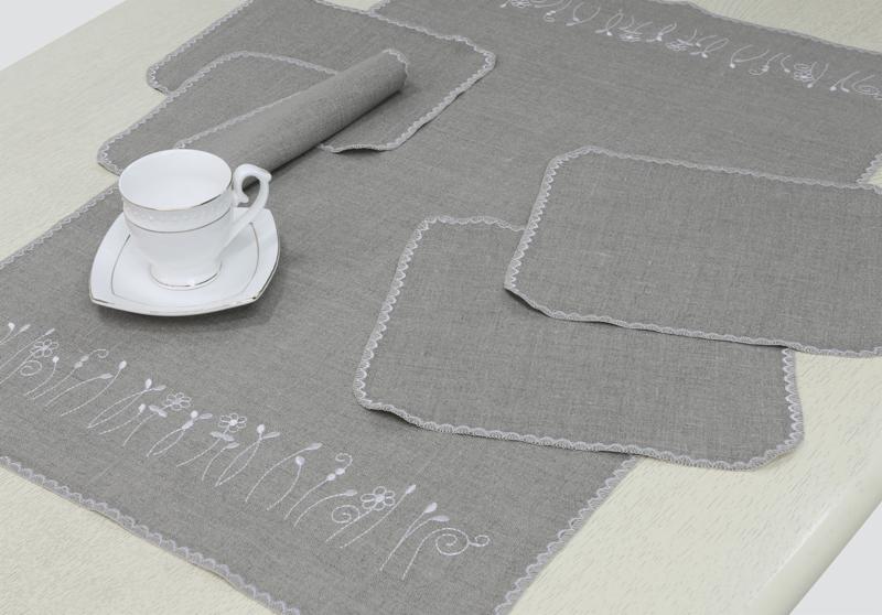 Lniany zestaw: bieżnik z haftem kwiatowym, podkładki z falbanką i zwinięta serwetka, uzupełniony filiżanką na białym stole.