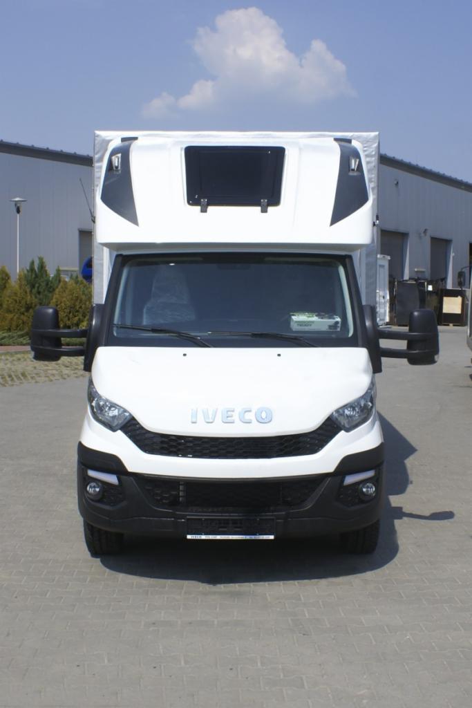 Biały samochód dostawczy marki Iveco z zabudową kontenerową, widok z przodu na tle hali magazynowej i błękitnego nieba z chmurami.