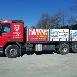 Czerwona ciężarówka z reklamą składu budowlanego LION I i logo firm Knauf, Wienerberger oraz Austrotherm, zaparkowana na żwirowym podłożu w słoneczny dzień.