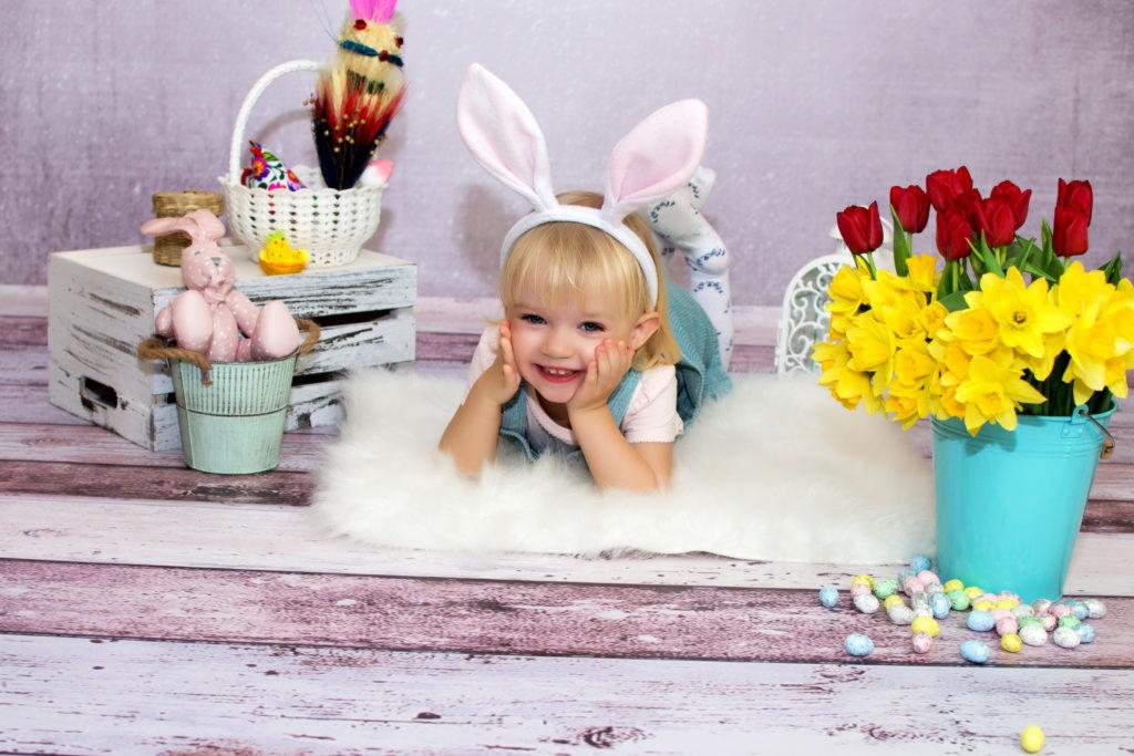 Uśmiechnięta dziewczynka z blond włosami i króliczymi uszami leży na białym futrze, opierając policzki na dłoniach, obok niej turkusowe wiadro z żółtymi narcyzami i czerwonymi tulipanami, a także...