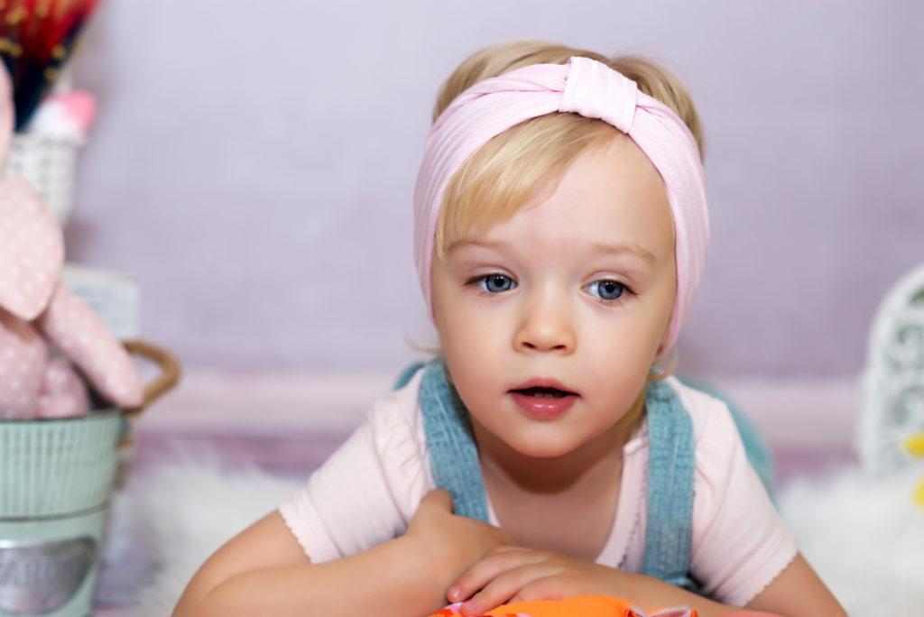 Portret małej dziewczynki z niebieskimi oczami i różową opaską na głowie, leżącej na białym futrze, z dekoracjami w tle.