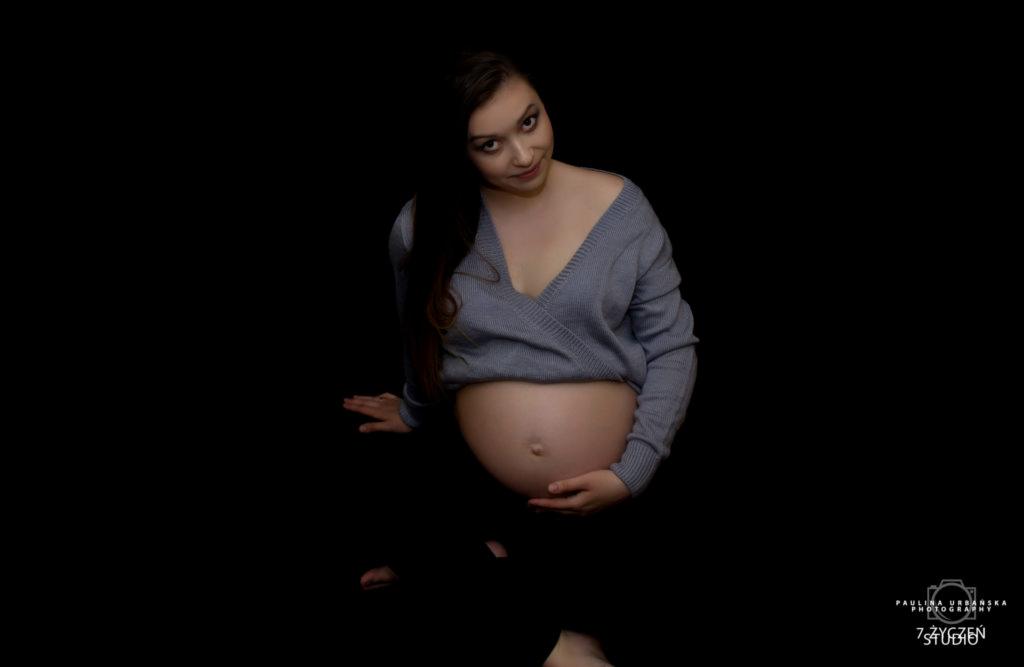 Kobieta w szarym swetrze z dekoltem w serek, trzymająca dłonie na ciążowym brzuchu, pozuje na czarnym tle, delikatny uśmiech.