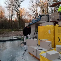 Pracownik w czarnej bluzie tnący bloczek z betonu komórkowego piłą elektryczną na budowie domu jednorodzinnego, w tle drugi pracownik na rusztowaniu i zapakowane bloczki YTONG.