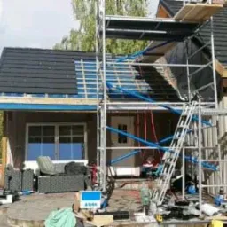 Dom jednorodzinny w trakcie prac dekarskich: częściowo ułożone czarne dachówki, rusztowanie z niebieskimi elementami, dekarz przy pracy na ziemi, widoczny fragment ogrodu i samochód dostawczy.