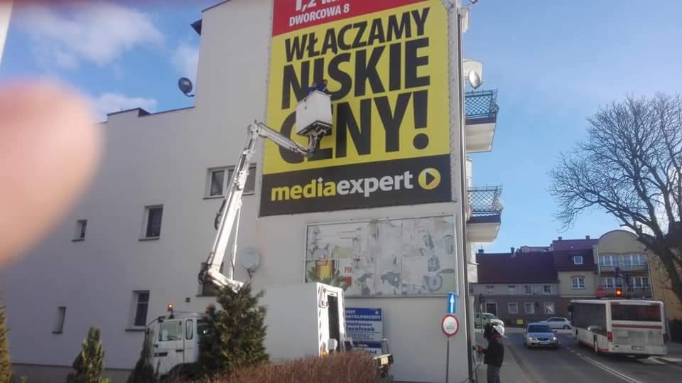 Podnośnik koszowy na tle budynku z reklamą Media Expert, pracownik w kasku na chodniku, widoczny autobus miejski i drzewo bez liści.