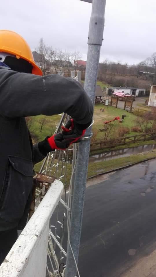 Pracownik w pomarańczowym kasku i czerwono-czarnych rękawicach montuje oświetlenie na metalowym słupie, widoczne szczypce i przewody, w tle zabudowania i pochmurne niebo.
