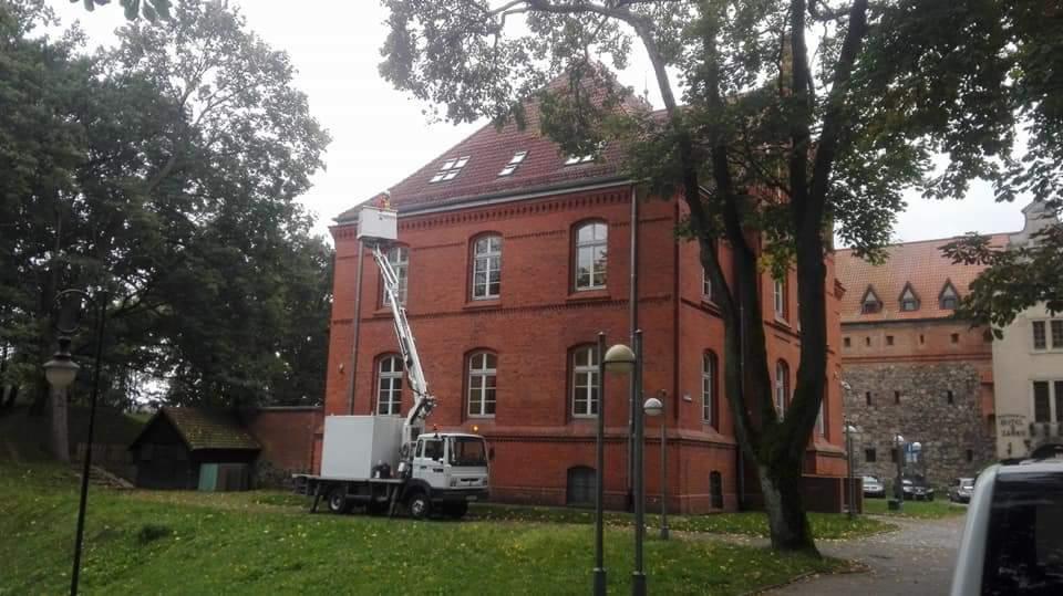 Ciężarówka z podnośnikiem koszowym zaparkowana przed ceglanym budynkiem z czerwonym dachem i białymi ramami okiennymi. W koszu podnośnika widoczna sylwetka osoby. W tle drzewa i fragmenty innych...