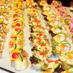 Rzędy kolorowych kanapek na białym pieczywie, z różnorodnymi dodatkami warzywnymi i dekoracjami, przygotowane przez firmę z Byczyny.