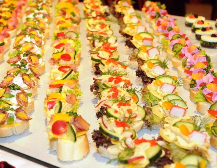 Rzędy kolorowych kanapek na białym pieczywie, z różnorodnymi dodatkami warzywnymi i dekoracjami, przygotowane przez firmę z Byczyny.