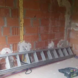 Wnętrze w trakcie prac: murowana ściana z instalacjami elektrycznymi w peszlu, aluminiowa drabina oparta o ścianę, skrzynka narzędziowa na podłodze.
