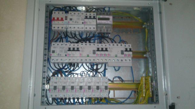 Rozdzielnica elektryczna z zainstalowanymi bezpiecznikami, licznikiem i okablowaniem w skrzynce metalowej, widok z góry.