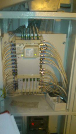 Rozdzielnica elektryczna w metalowej obudowie z licznymi kablami podłączonymi do złączek, widoczne diody sygnalizacyjne.