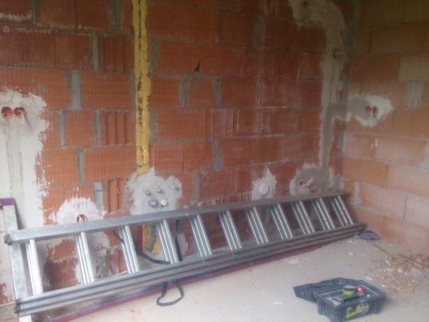 Wnętrze w trakcie prac: murowana ściana z instalacjami elektrycznymi w peszlu, aluminiowa drabina oparta o ścianę, skrzynka narzędziowa na podłodze.