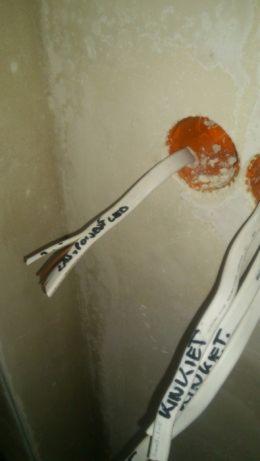 Fragment ściany z dwoma otworami instalacyjnymi, z których wystają przewody elektryczne z ręcznie naniesionymi napisami, w tym '230V faza LED' i 'KINKIET KINKIET'.