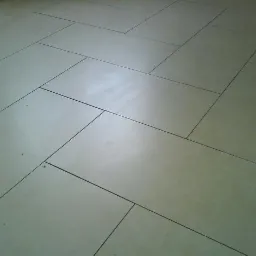 Beżowe, prostokątne płytki ułożone w jodełkę na podłodze, widoczne delikatne zabrudzenia w fugach.