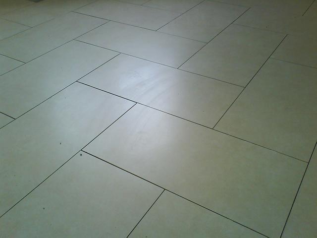 Beżowe, prostokątne płytki ułożone w jodełkę na podłodze, widoczne delikatne zabrudzenia w fugach.