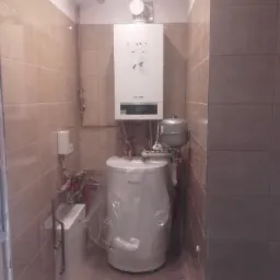 Biały kondensacyjny piec gazowy zamontowany nad podgrzewaczem wody, instalacja w łazience wykończonej beżowymi płytkami, podgrzewacz częściowo owinięty folią ochronną.