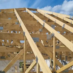 Konstrukcja drewniana więźby dachowej z widocznymi metalowymi łącznikami na tle błękitnego nieba z chmurami.