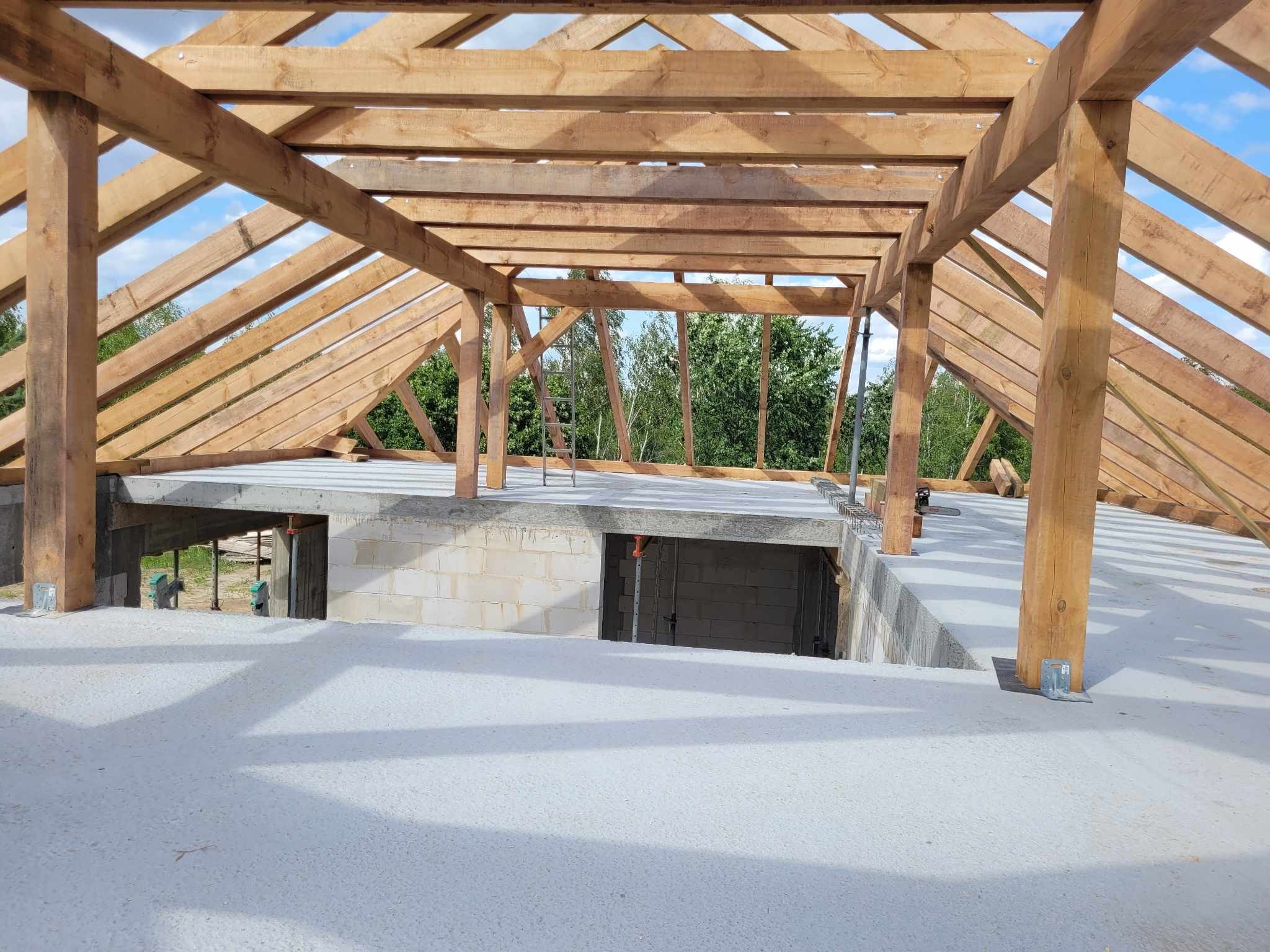 Konstrukcja drewniana więźby dachowej w trakcie budowy, widok od wewnątrz budynku, z betonu wylany strop, na zewnątrz widoczne drzewa.