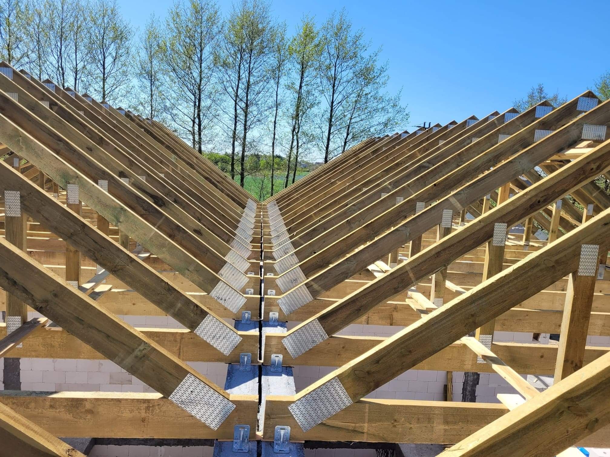 Konstrukcja drewniana dachu z widocznymi metalowymi łącznikami na tle błękitnego nieba i drzew