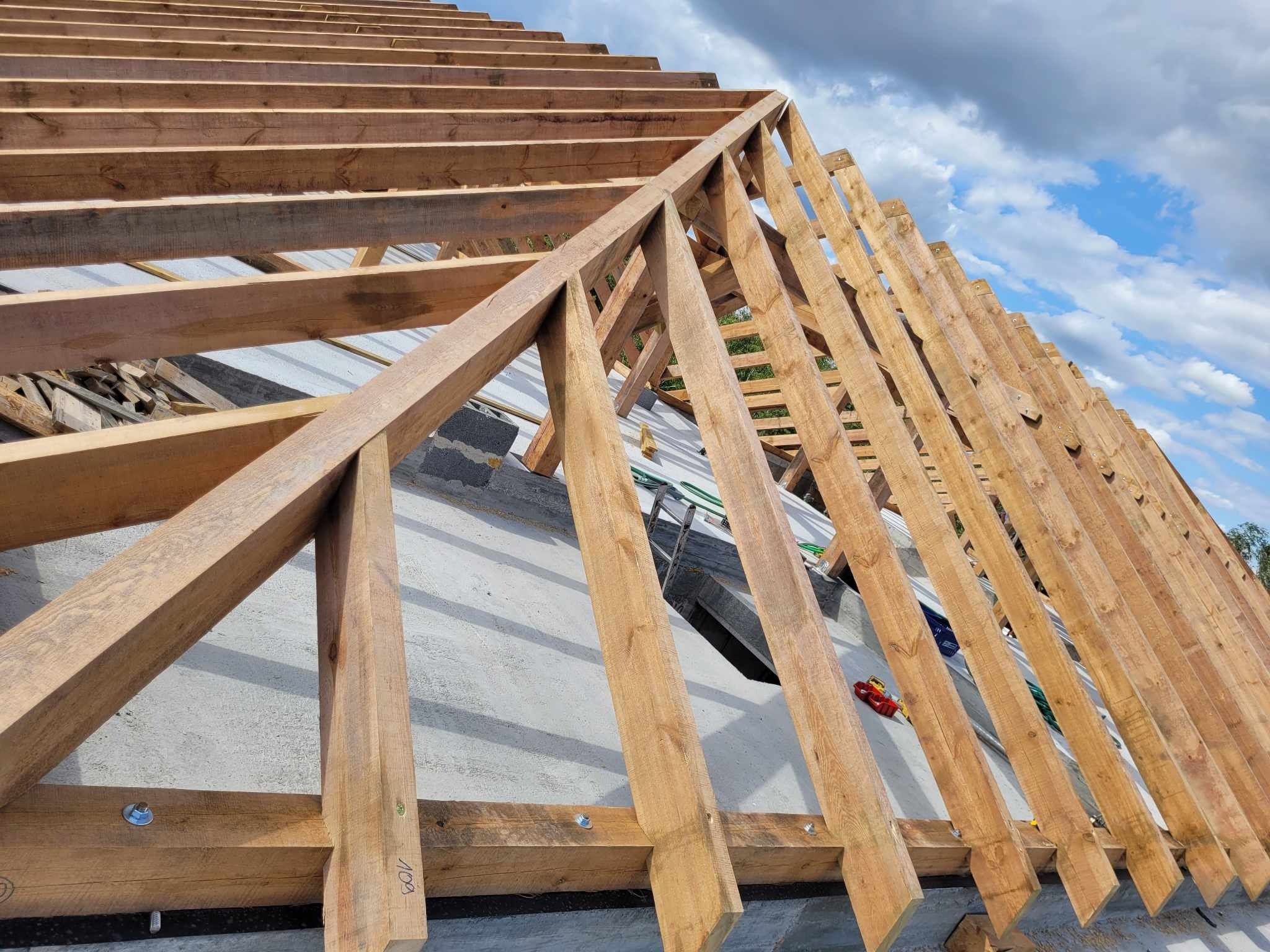 Konstrukcja drewniana więźby dachowej w trakcie montażu, widoczne elementy mocowane śrubami, na tle błękitnego nieba z chmurami.