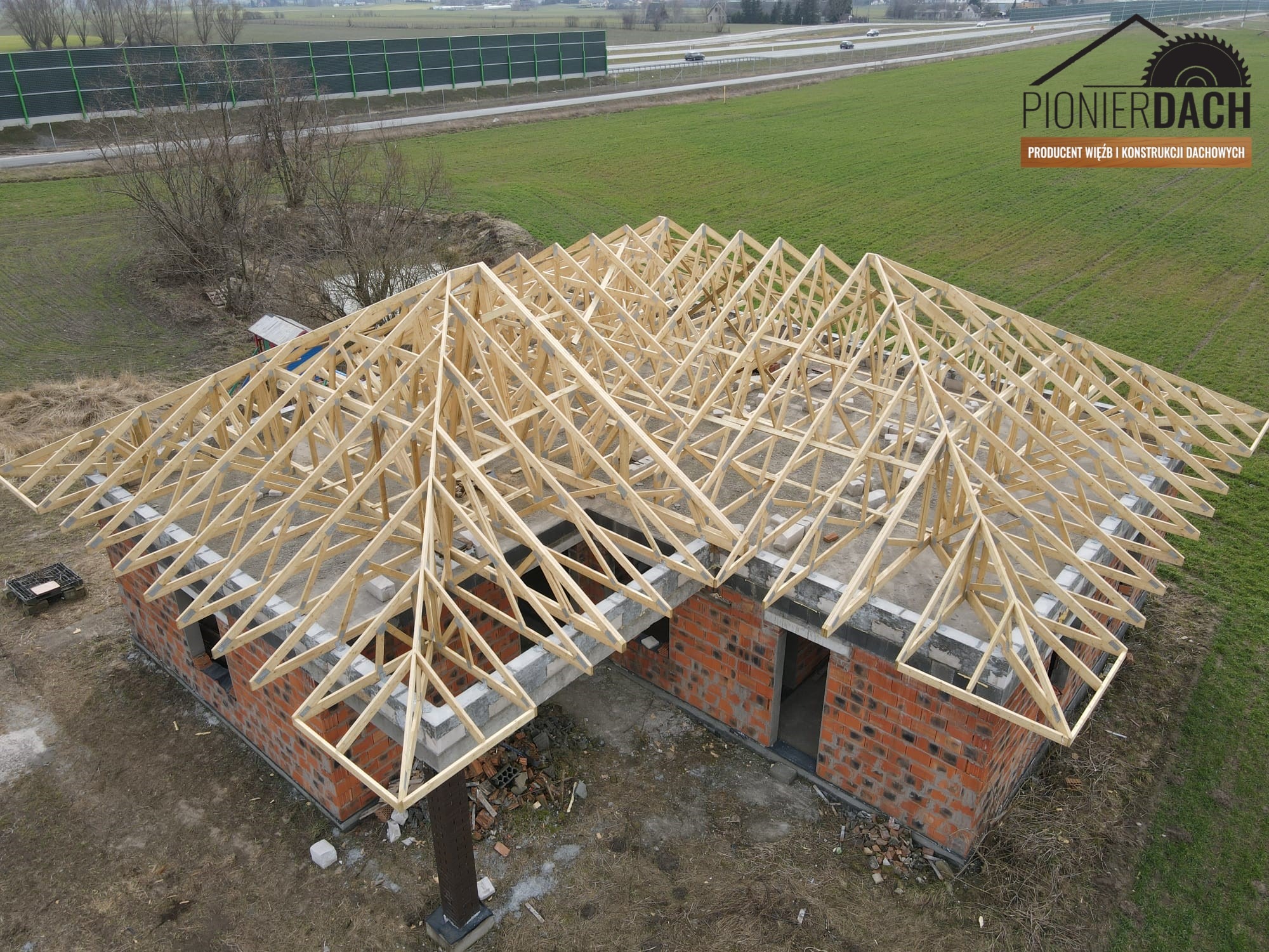 Wiązary dachowe z jasnego drewna na murowanym domu w Pruszczu, widok z góry, w tle droga szybkiego ruchu i logo firmy.