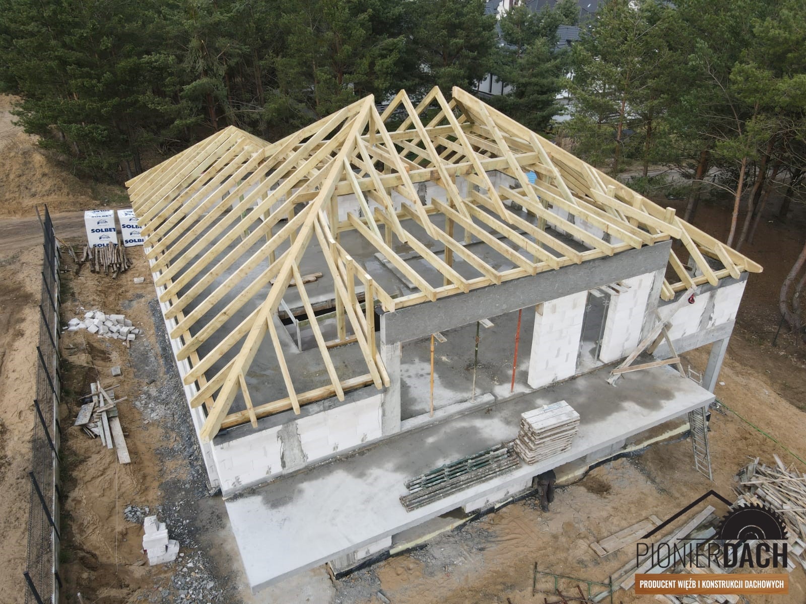 Wiązary dachowe z drewna na murowanym budynku w stanie surowym otwartym, widok z góry, w tle las iglasty i logo firmy dekarskiej w prawym dolnym rogu.