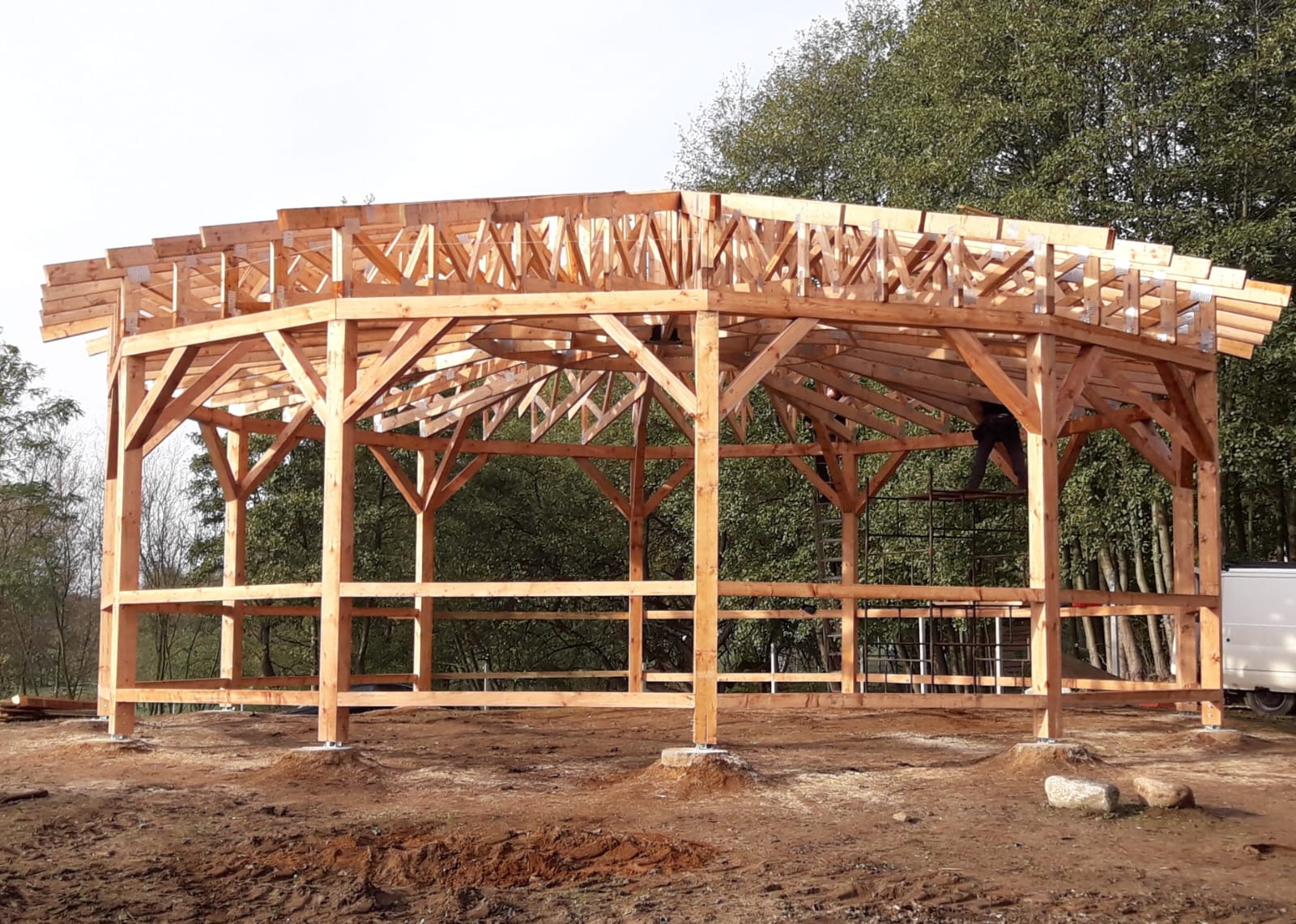 Konstrukcja drewniana dachu w trakcie budowy, widoczne elementy więźby i słupy nośne, zarys sylwetki pracownika na rusztowaniu w tle.