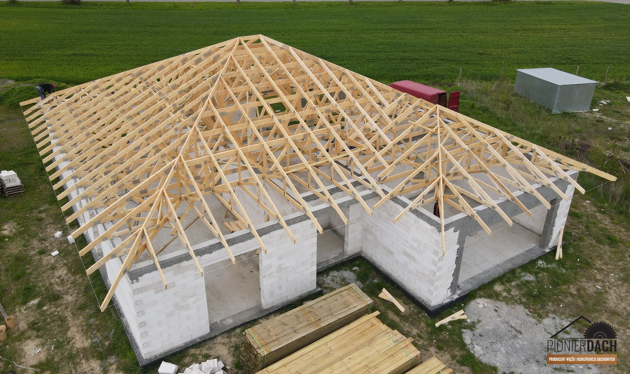 Wznoszenie więźby dachowej na budynku z betonu komórkowego, widok z góry na konstrukcję z jasnego drewna, w tle zielone pole i czerwony samochód dostawczy.