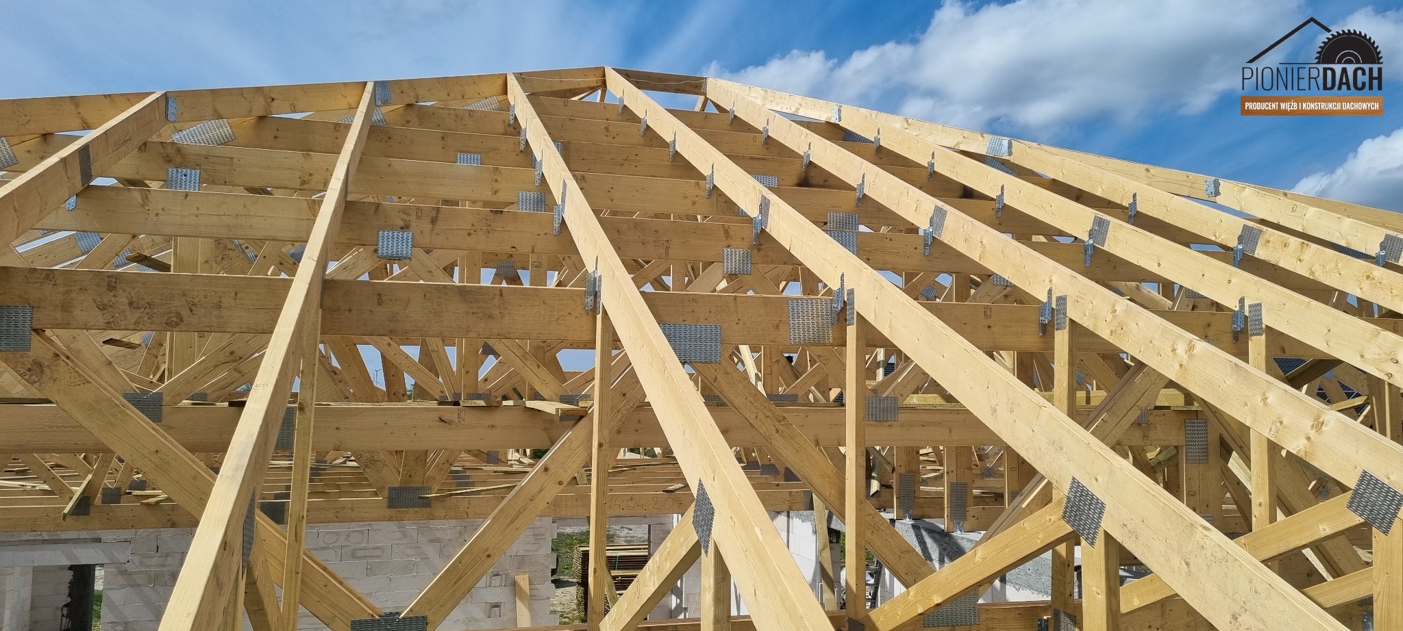 Konstrukcja drewniana więźby dachowej z widocznymi metalowymi łącznikami na tle błękitnego nieba z chmurami.