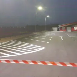 Nowo wyznaczone, białe pasy na parkingu przed czerwonym budynkiem, oświetlone przez latarnie uliczne, z taśmą ostrzegawczą w poprzek kadru.