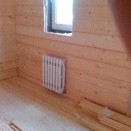 Maksim Halushka MaxiSoft - Wnętrze domu w trakcie wykańczania z drewnianą boazerią, zamontowany biały grzejnik żeberkowy pod oknem, widoczne deski podłogowe i fragmenty materiałów budowlanych.