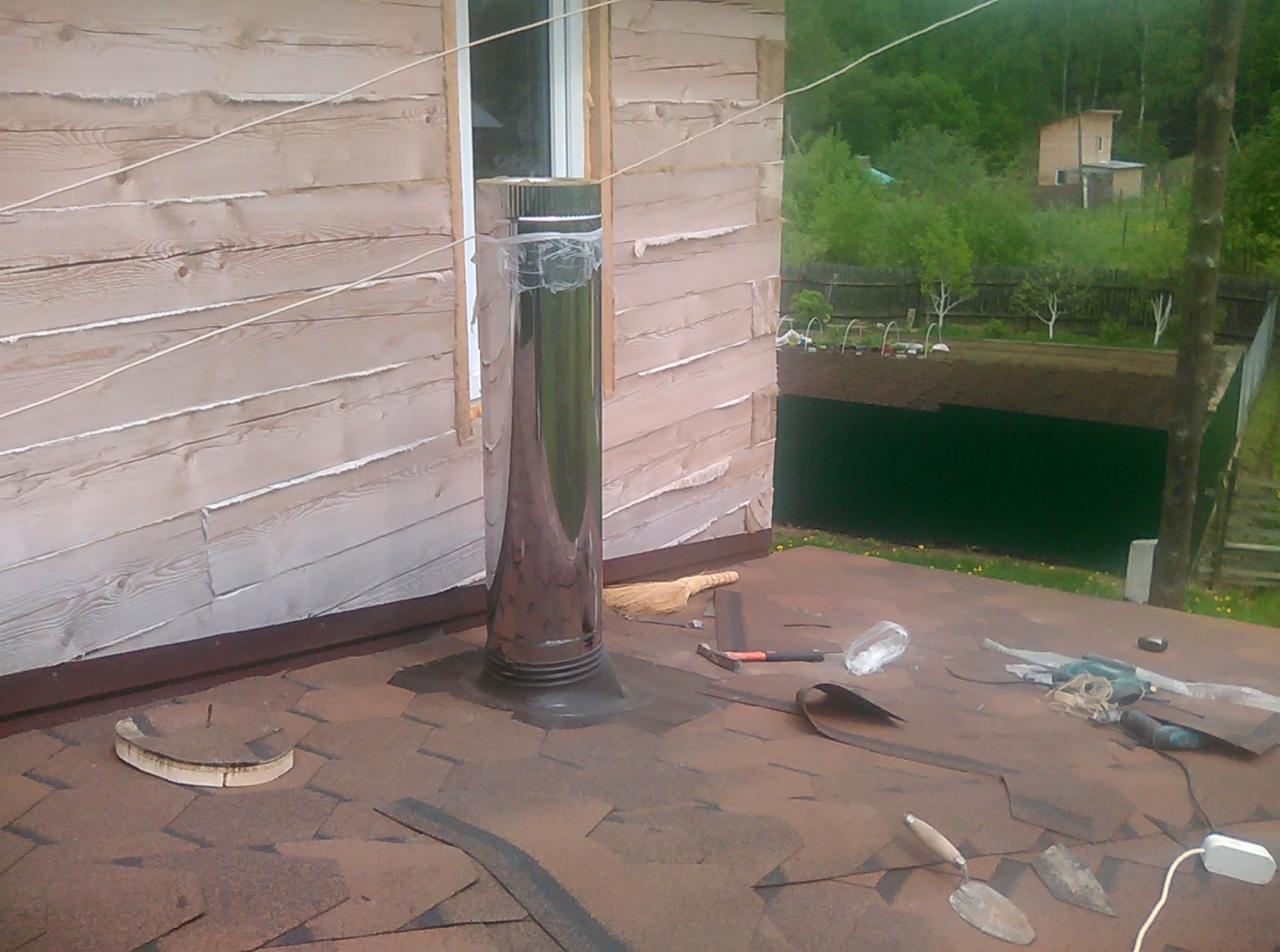 Montaż komina stalowego na dachu pokrytym gontem bitumicznym, widoczne narzędzia, częściowo zdjęta folia ochronna z komina, drewniana elewacja budynku w tle.