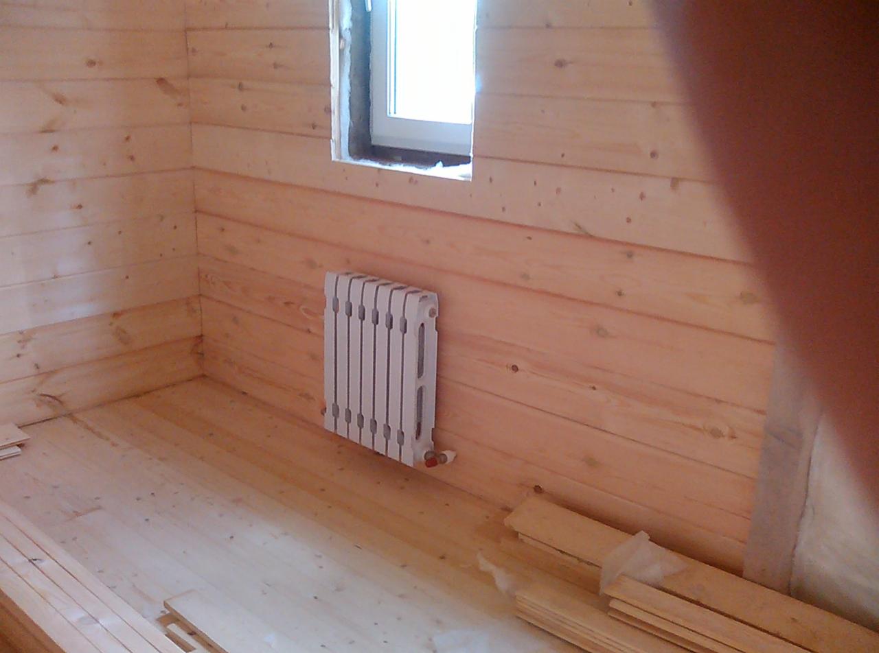 Wnętrze domu w trakcie wykańczania z drewnianą boazerią, zamontowany biały grzejnik żeberkowy pod oknem, widoczne deski podłogowe i fragmenty materiałów budowlanych.