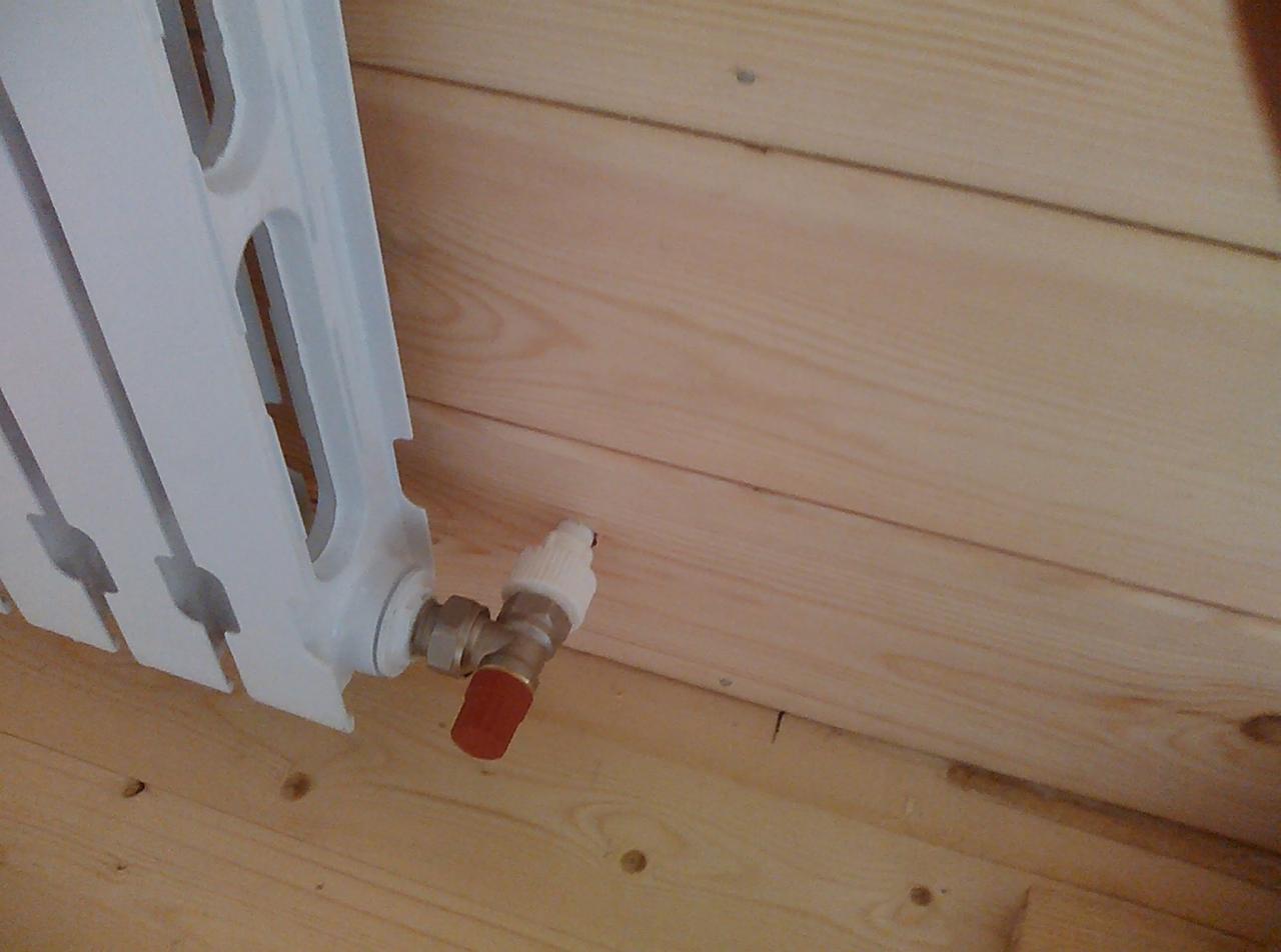 Biały, żeberkowy grzejnik zamontowany przy drewnianej ścianie i podłodze, z widocznym zaworem termostatycznym z czerwoną nakładką.