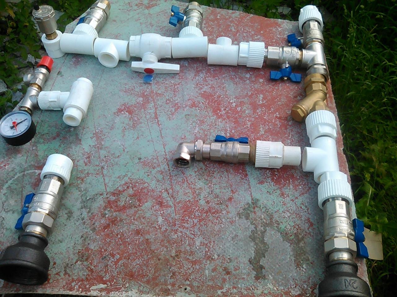Rozłożone na czerwonej, popękanej powierzchni elementy instalacji wodnej: białe rury PCV, metalowe złączki z niebieskimi zaworami, manometr z czerwoną nakrętką, widoczne trawa i krzewy w tle.