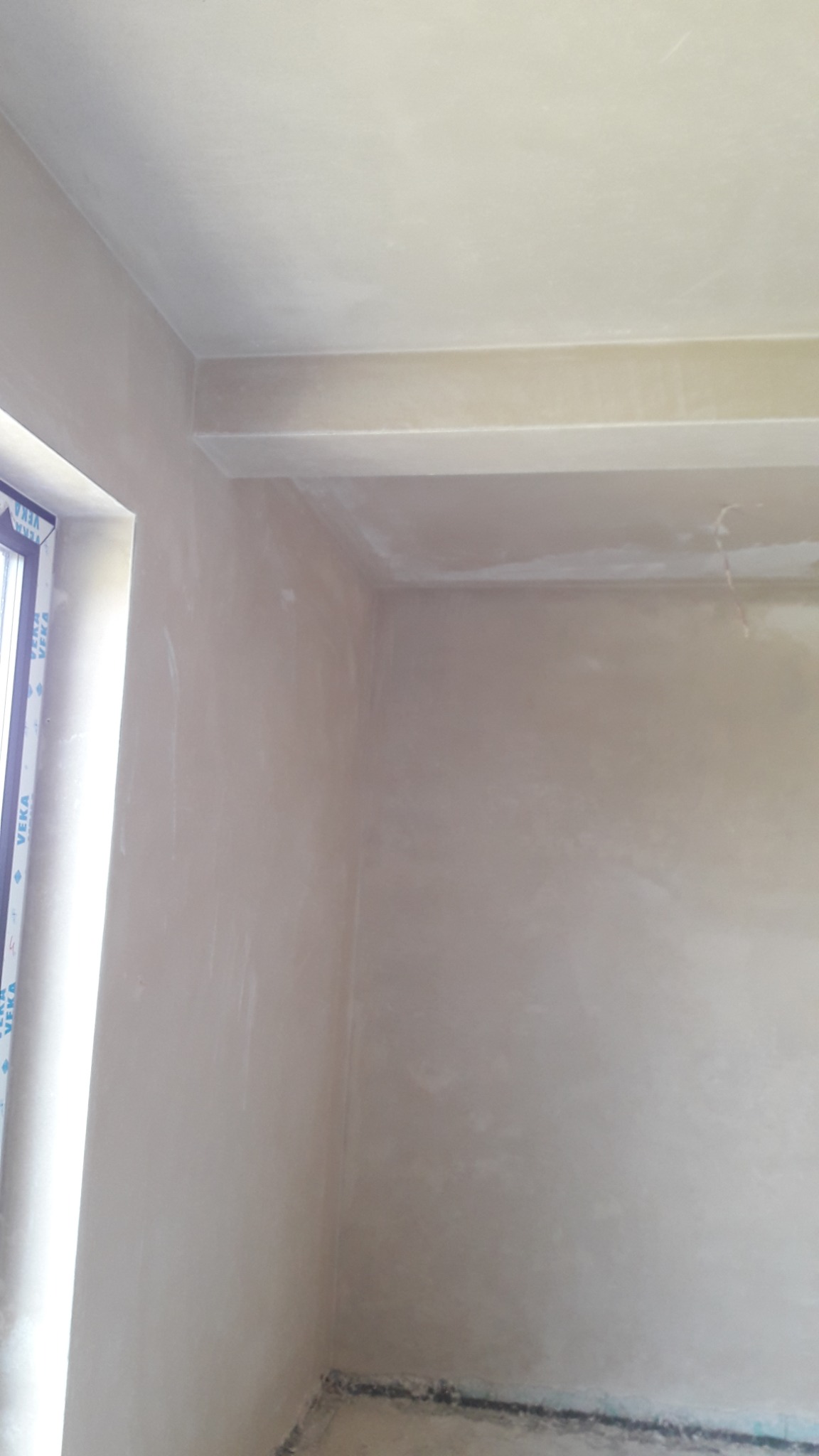 Wnętrze pokoju po nałożeniu świeżej, jasnej warstwy gładzi szpachlowej na ścianach i suficie, widoczne delikatne nierówności i ślady po narzędziach, okno po lewej stronie kadru.