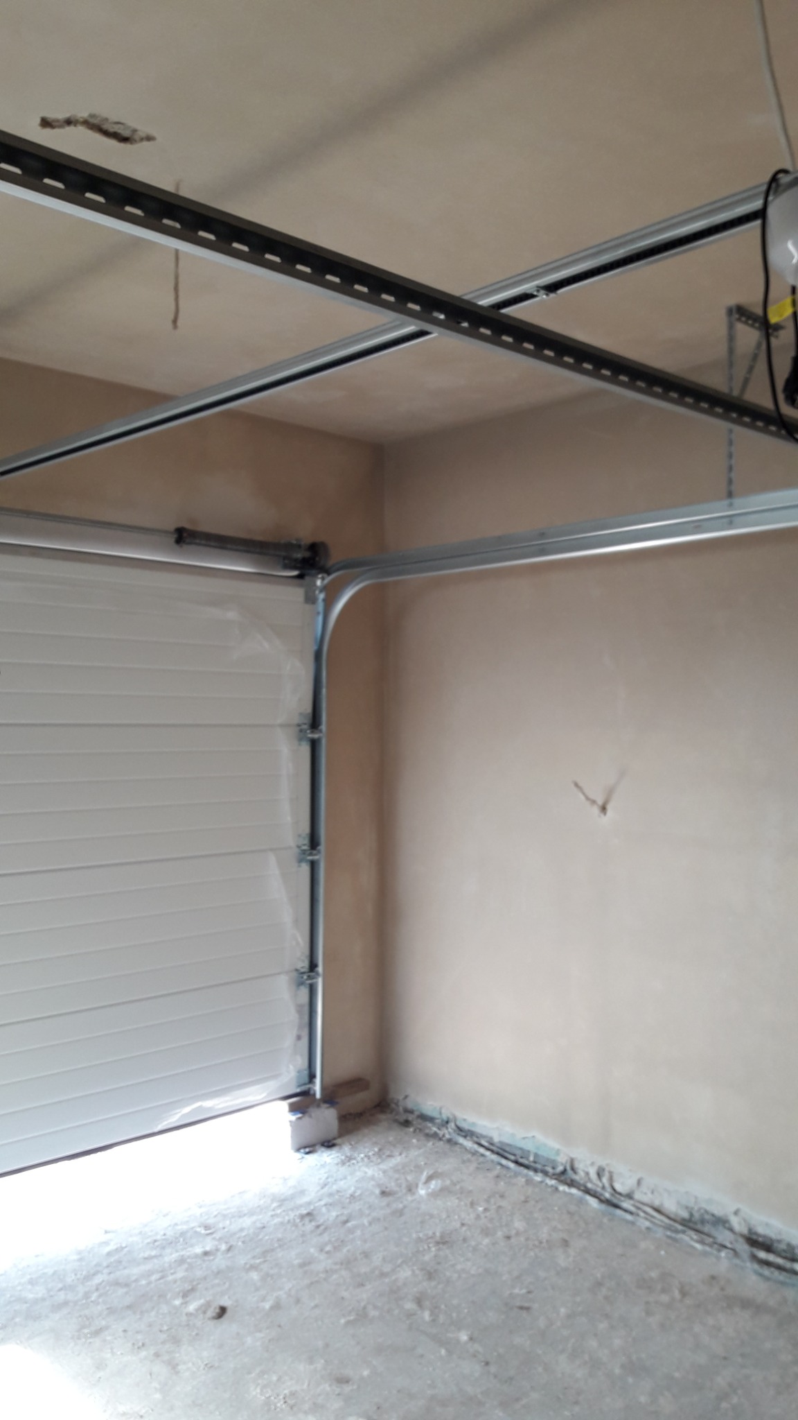 Wnętrze garażu w trakcie prac wykończeniowych: widoczne szyny do bramy garażowej, surowa ściana i betonowa podłoga przed ostatecznym wykończeniem.