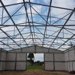 Konstrukcja stalowa hali w trakcie budowy, widoczna szkieletowa konstrukcja dachu i ściany z bloczków betonowych.