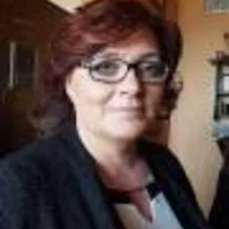 Portret kobiety w okularach, z krótkimi rudymi włosami, ubranej w ciemną marynarkę i białą bluzkę, w neutralnym wnętrzu.