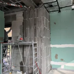 Wnętrze w trakcie wykańczania: montaż płyt gipsowo-kartonowych na ścianie, widoczne metalowe profile sufitowe, fragment ściany z surowego betonu i pracownik na rusztowaniu.