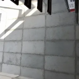 Szara, betonowa podłoga ułożona z prostokątnych płyt, oświetlona promieniami słońca tworzącymi geometryczne cienie, widoczne elementy konstrukcji dachu i paczka materiałów budowlanych w rogu.