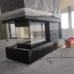 Nowoczesny, przeszklony kominek narożny z czarną obudową, częściowo zakryty folią ochronną w trakcie prac wykończeniowych, widoczny kubeł budowlany w tle.