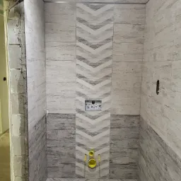 Wnętrze łazienki w trakcie remontu, z ułożonymi płytkami imitującymi drewno w odcieniach szarości i bieli, wzór w jodełkę na podłodze i pas z motywem strzałek na ścianie, widoczne elementy...