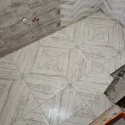 Ułożone płytki podłogowe i ścienne imitujące drewno w łazience, wzór geometryczny na podłodze, widoczne przyłącza wodne i pojemnik z zaprawą, etap prac wykończeniowych.