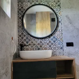 Nowoczesna łazienka w Orzeszu z okrągłym lustrem, nablatową umywalką i szafką z drewnianym blatem oraz dekoracyjnymi płytkami na ścianie, widoczne przewody wystające ze ściany po lewej stronie.