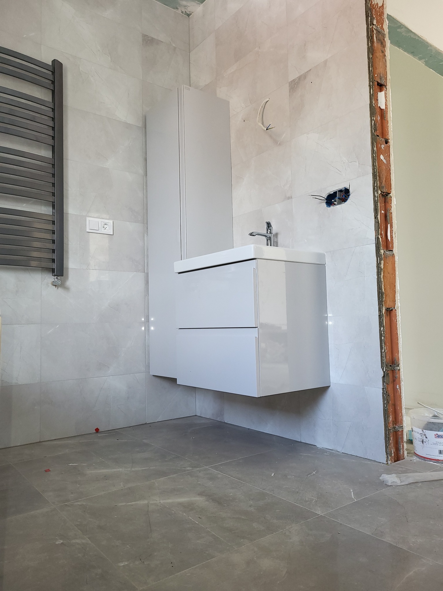 Montaż umywalki z szafką w łazience wyłożonej płytkami imitującymi marmur, widoczne niedokończone obramowanie drzwi oraz grzejnik drabinkowy.