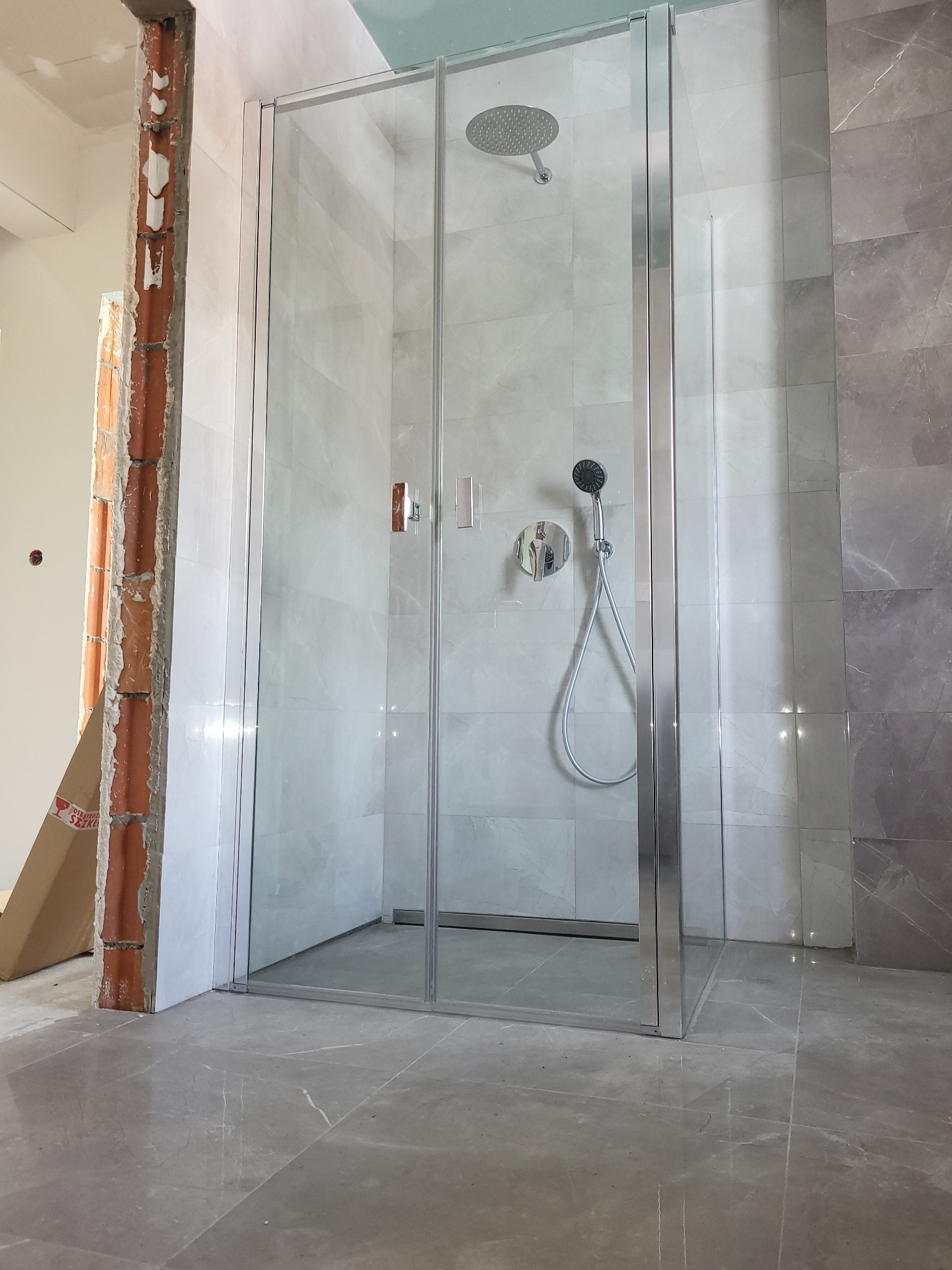 Nowoczesna kabina prysznicowa z szarymi płytkami, widoczny fragment ściany w stanie surowym i kartonowe pudło, sugerujące trwający remont.