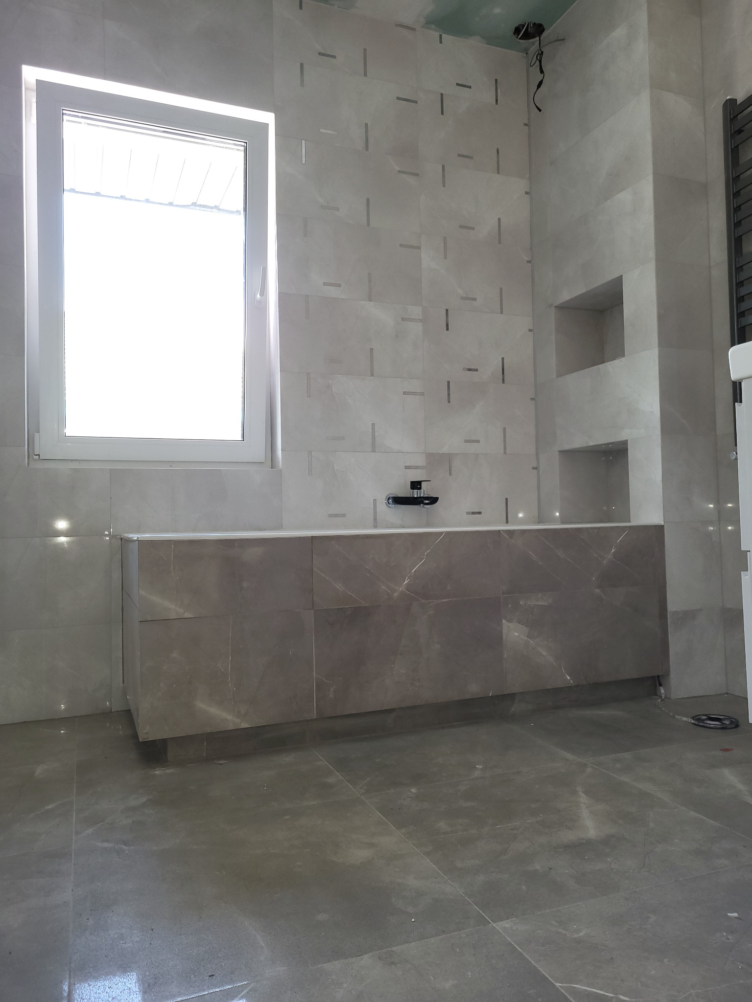 Jasna łazienka w trakcie wykańczania, z szarymi płytkami imitującymi marmur na ścianach i podłodze, widoczna zabudowa umywalkowa z ciemniejszego marmuru, okno i wnęki w ścianie, sufit częściowo...